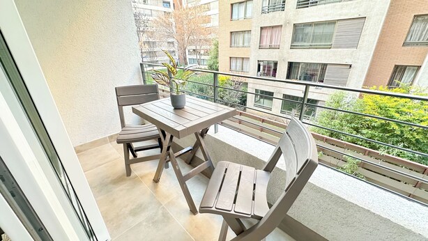 A balcony with a table and chairs, a potted plant, and a view of a building with multiple windows. - Moderno depto. en Stgo. cerca del metro (Santiago)