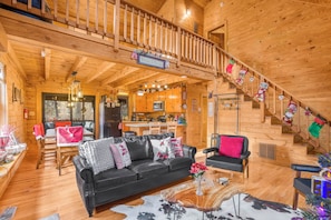 A wooden cabin interior with a black leather sofa, a dining area, a kitchen, and a staircase.