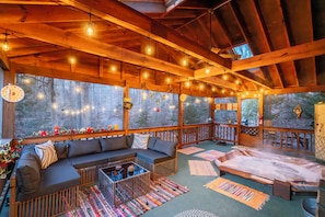A cozy cabin interior with a wooden ceiling, string lights, a sofa, and a view of the forest.