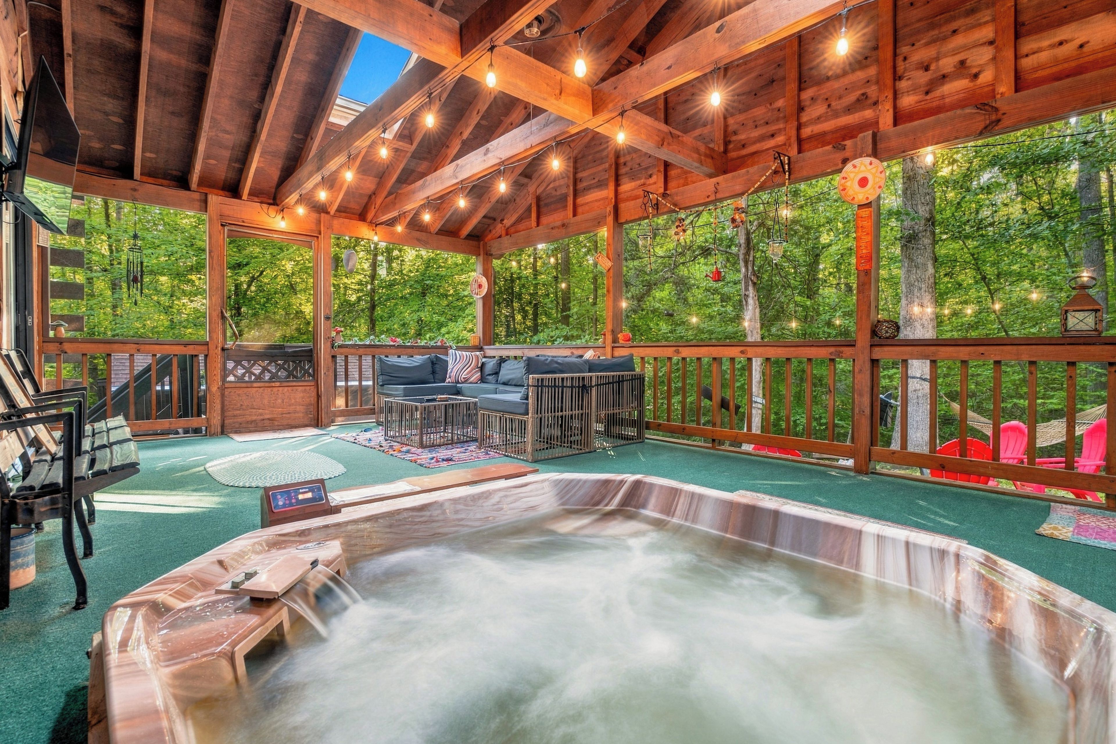 A covered outdoor area with a hot tub, seating, and a view of the forest.