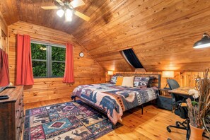 A cozy cabin bedroom with a wooden ceiling, a bed with a patterned quilt, a desk, and a chair.