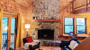 A cozy cabin interior with a stone fireplace, wooden walls, a comfortable seating area, and a window overlooking the forest.
