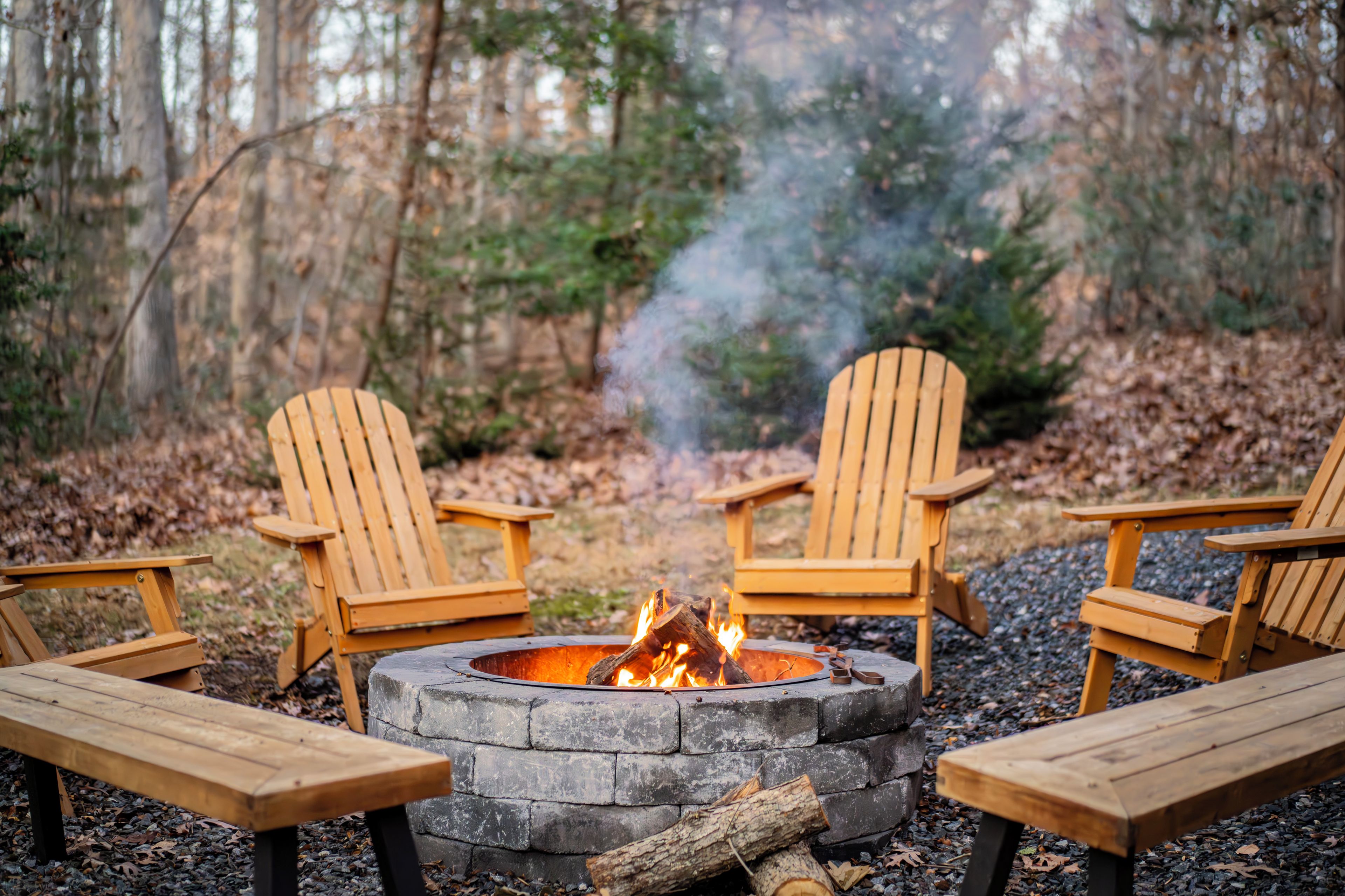 A campfire with four wooden Adirondack chairs around it, set in a forested area with a stone fire pit.
