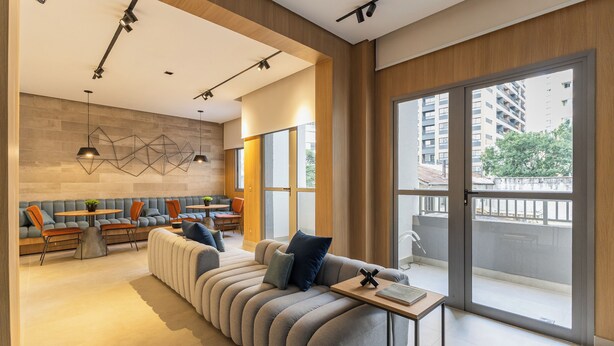 A modern living room with a sofa, chairs, a table, and a large window with a view of buildings and trees. - Stay Home João Cachoeira - Piscina - Gym (São Paulo)