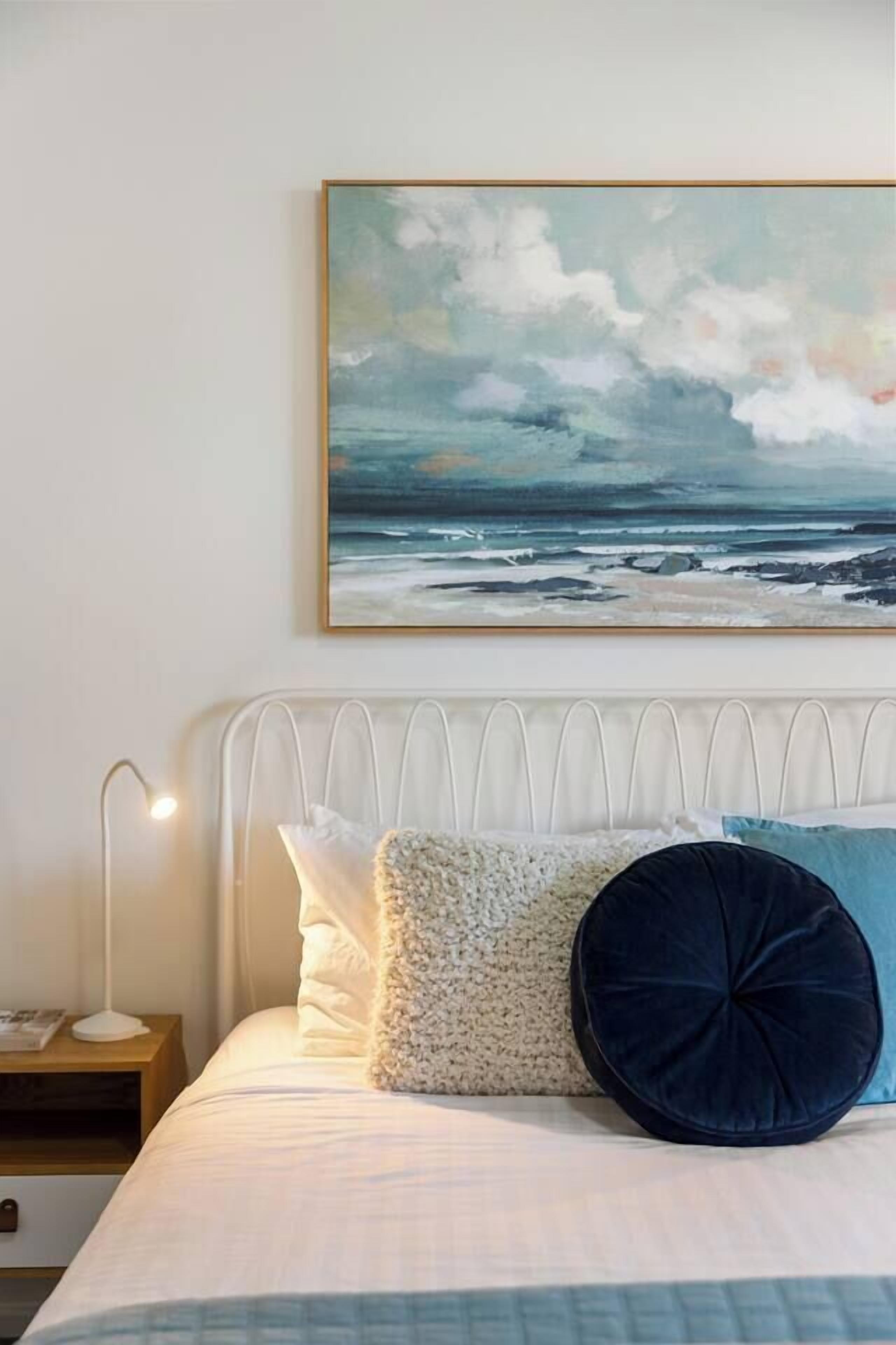 A bedroom with a bed, pillows, a bedside table, a lamp, and a painting of a beach scene.