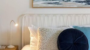 Een slaapkamer met een bed, kussens, een nachtkastje, een lamp en een schilderij van een strand.
