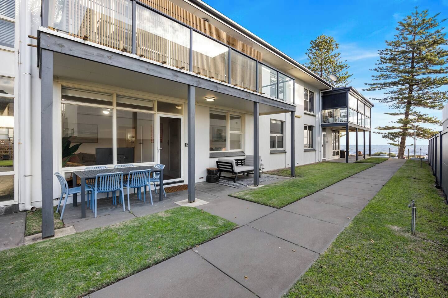 A modern two-story building with a covered patio, a dining area with blue chairs, and a view of the sea.