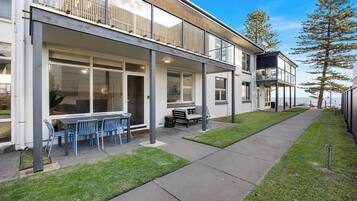 A modern two-story building with a covered patio, a dining area with blue chairs, and a view of the sea.