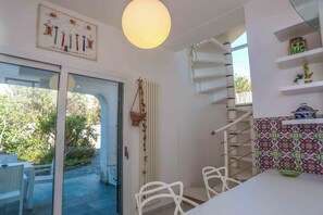 Habitació amb taula blanca, cadires, cistella penjant, radiador i paret amb taulells de colors.
