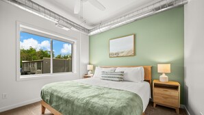 Een slaapkamer met een groot raam, een bed met een groene deken, twee nachtkastjes met lampen en een lijst met een afbeelding aan de muur.