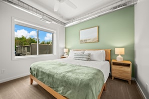 Ein Schlafzimmer mit großem Fenster, einem Bett mit grüner Decke, zwei Nachttischen mit Lampen und einem gerahmten Bild an der Wand.