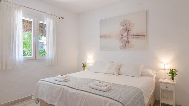 A bedroom with a bed, a bedside table with a lamp, a window with curtains, and a painting on the wall. - La Soberana: The House That Cares for You (Arroyo)