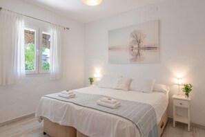 A bedroom with a bed, a bedside table with a lamp, a window with curtains, and a painting on the wall.
