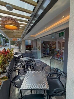 An outdoor cafe with tables and chairs, a glass door entrance, and a view of a store. - HOTEL AIKOS (Yumbo)