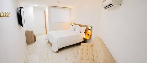 A hotel room with a bed, a sofa, a wall-mounted air conditioner, and a door leading to another room.