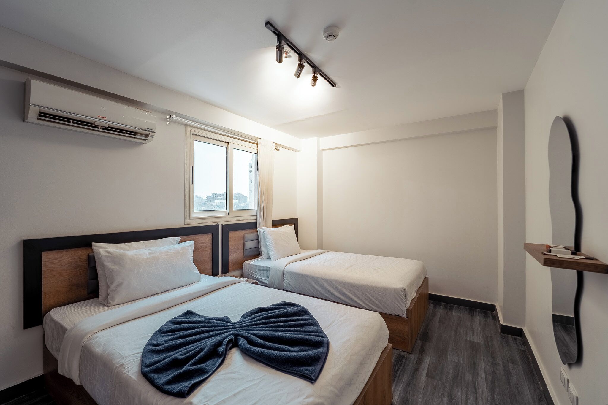 A hotel room with two beds, an air conditioning unit, a window, a mirror, and a ceiling light fixture.