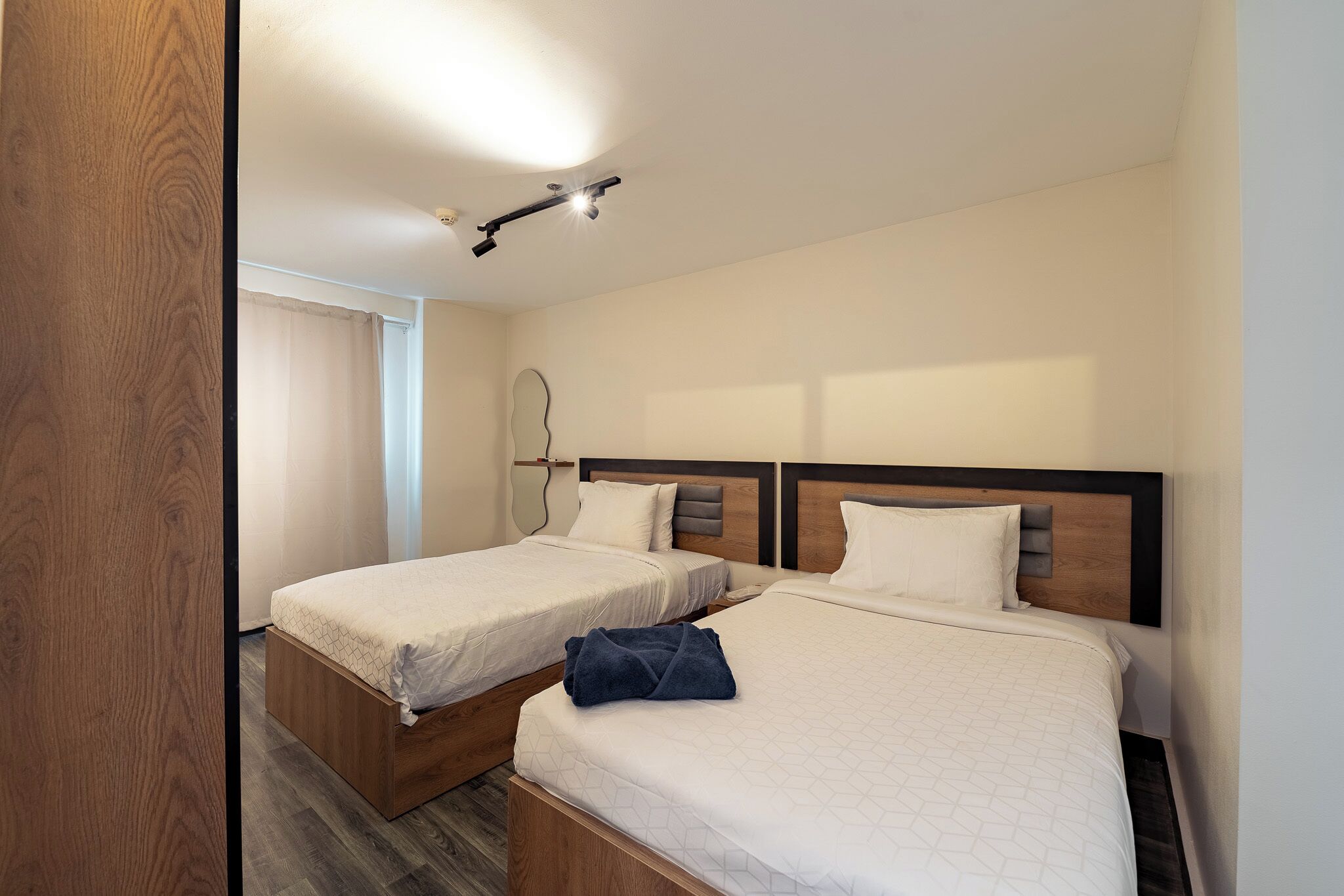 A hotel room with two beds, a wooden floor, a ceiling light, and a window with curtains.