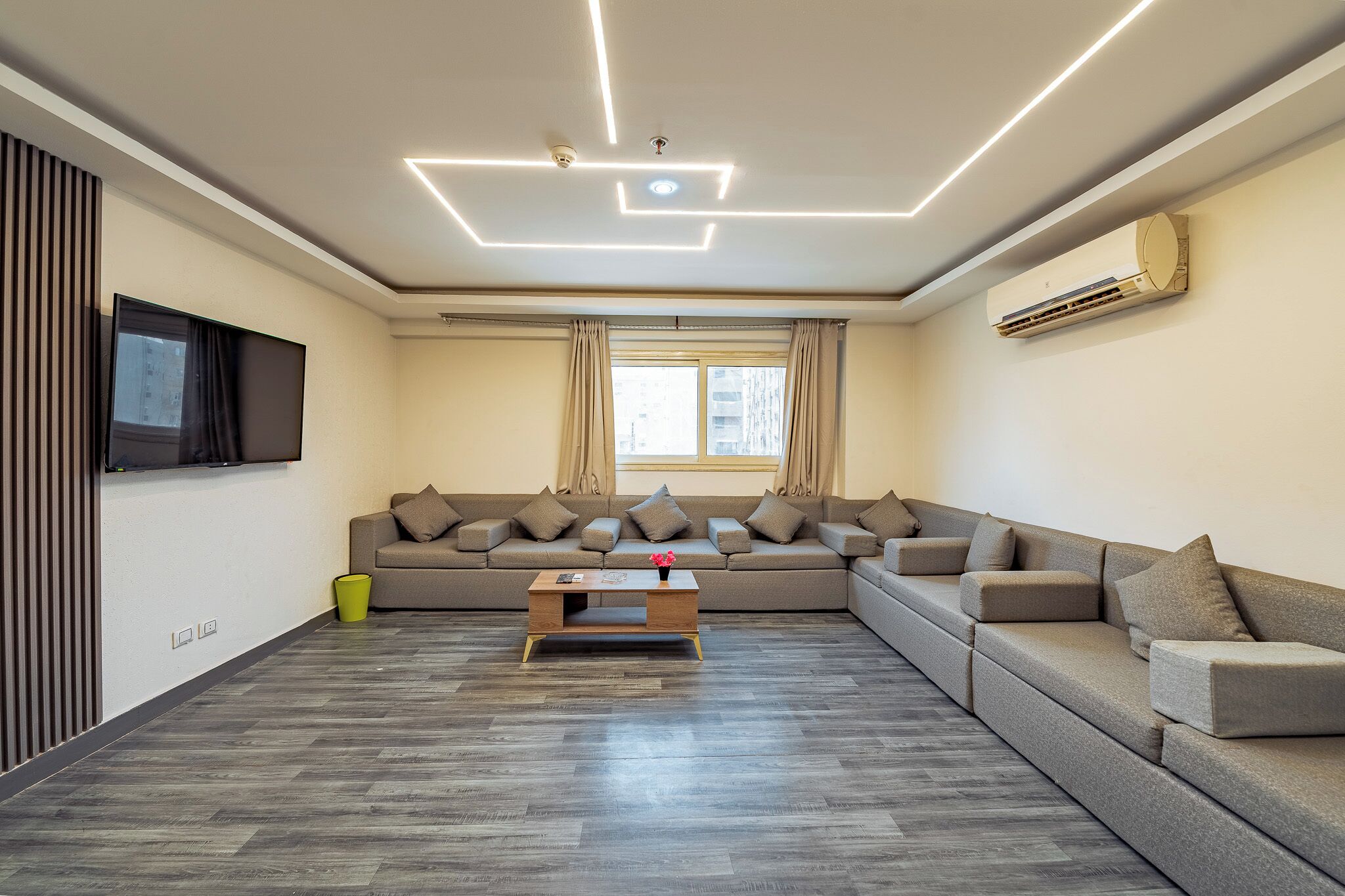 Ein modernes Wohnzimmer mit einem Ecksofa, einem Flachbildfernseher und einer Decke mit geometrischer Beleuchtung.