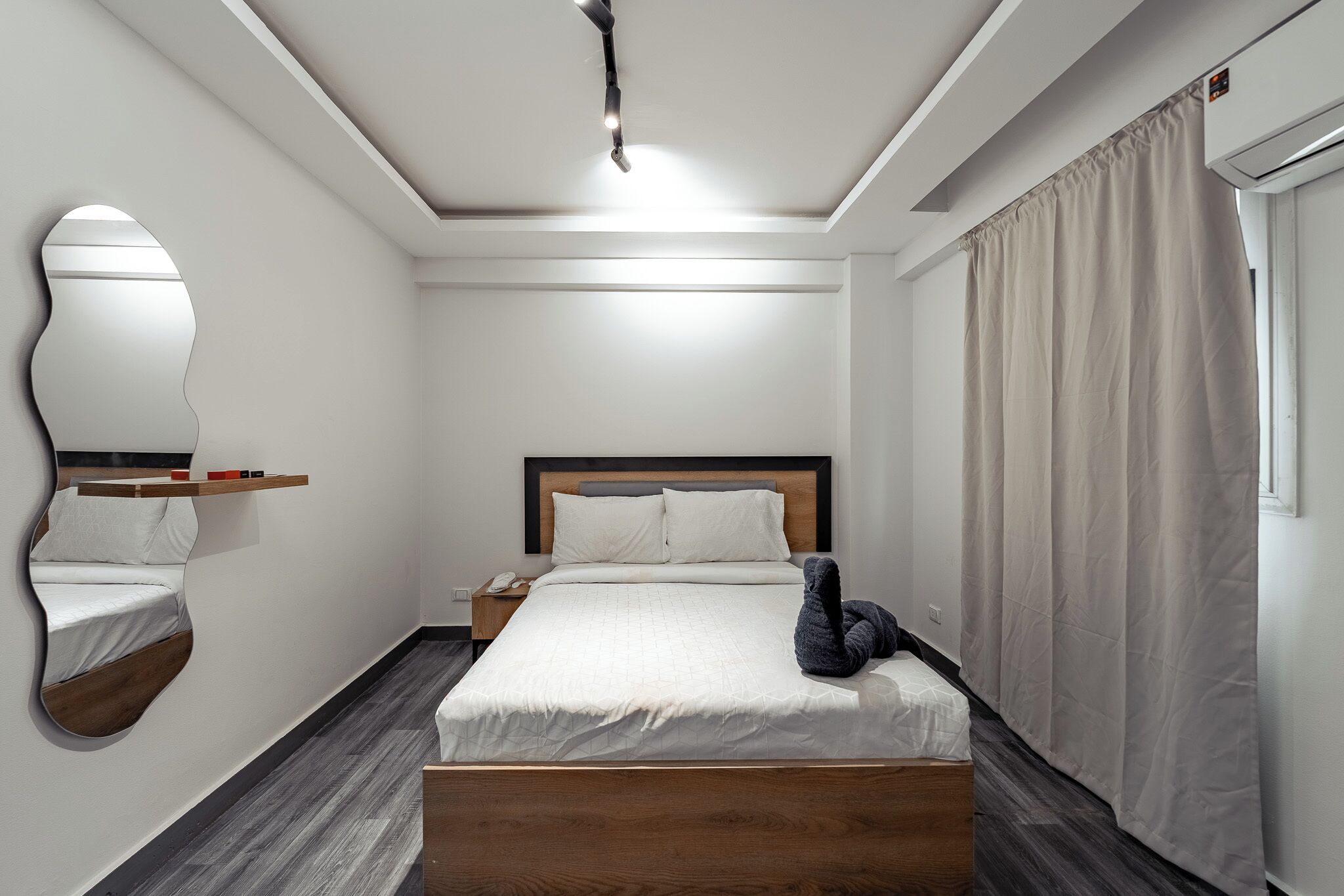 A modern hotel room with a wooden bed, a nightstand, a mirror, and a window with curtains.