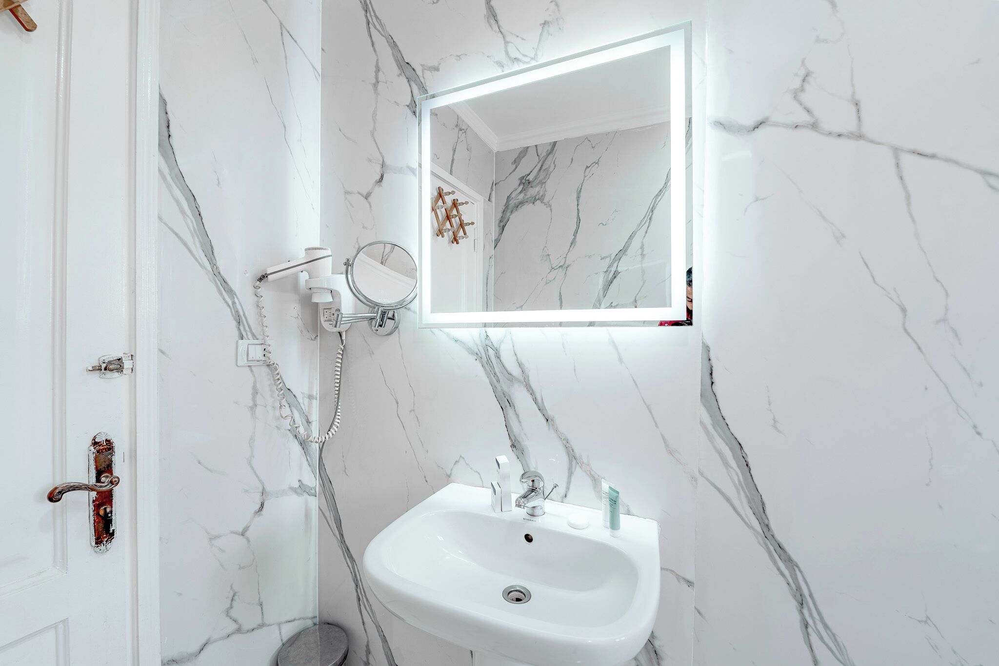 Un baño con lavabo de mármol blanco, ducha de mano, espejo con luz y puerta con pomo.