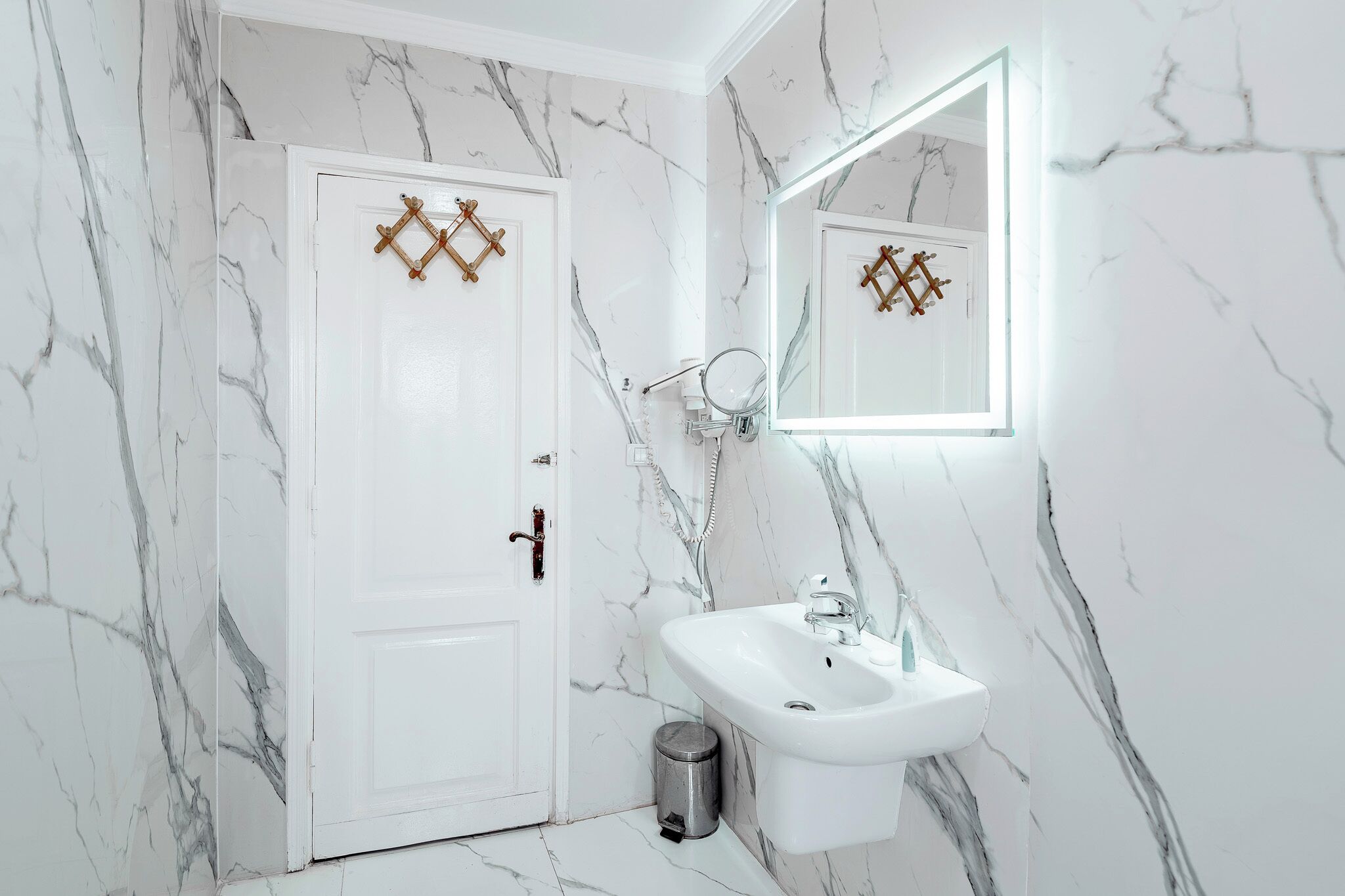 A bathroom with a white door, a sink, a mirror, and a trash bin.