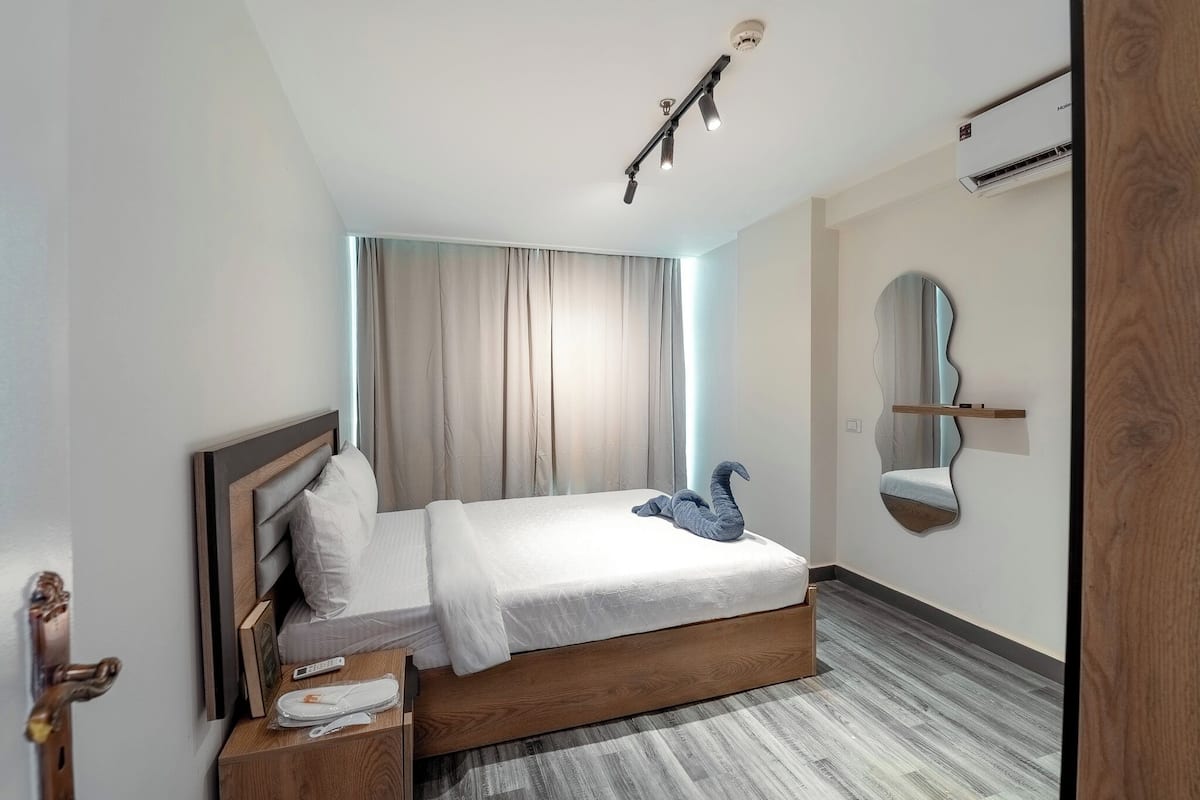 A modern hotel room with a wooden bed, a bedside table, a mirror, and an air conditioning unit.