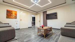 Una habitación de hotel moderna con un sofá gris, una mesa de centro de madera con un florero, un cuadro y un televisor de pantalla plana montado en la pared.