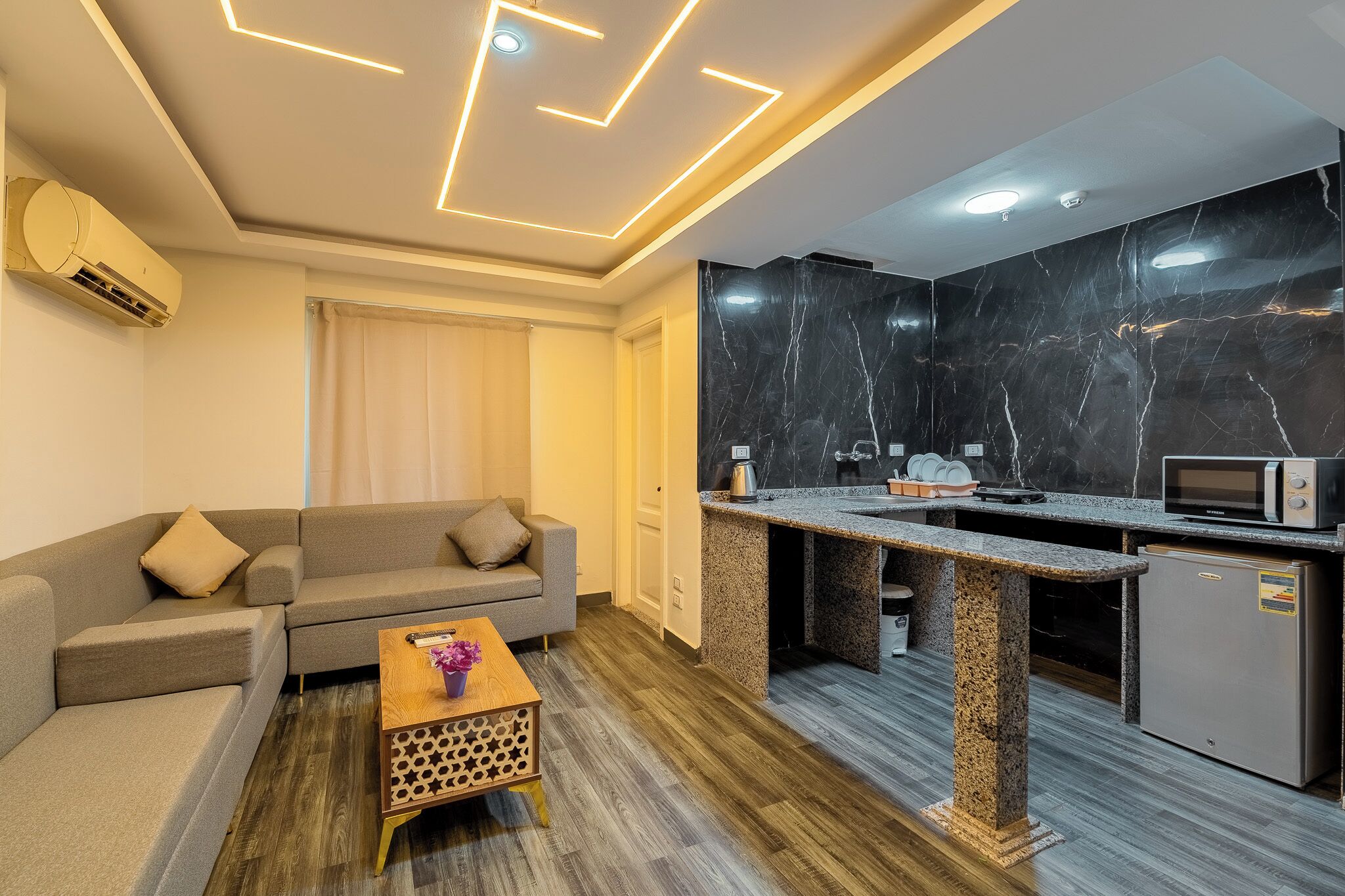 A modern living room with a sofa, a wooden coffee table, and a kitchen area with a marble backsplash and built-in appliances.