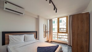 Una habitación de hotel con cama, unidad de aire acondicionado, ventana con cortinas y vista a edificios.