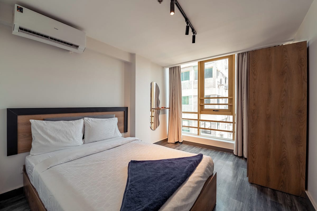 A hotel room with a bed, an air conditioning unit, a window with curtains, and a view of buildings outside.
