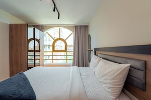 A hotel room with a large bed, a wooden headboard, a view through a window with curtains, and a modern light fixture.