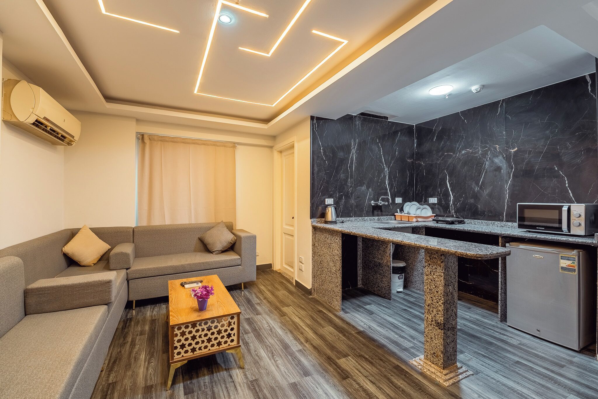 Een moderne kamer met een bank, een klein houten tafeltje, een kitchenette met een magnetron en een marmeren spatwand.