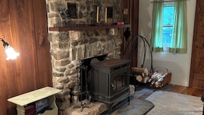 Una chimenea de piedra con repisa de madera, una mesita con libros, una alfombra estampada y una ventana con cortinas.