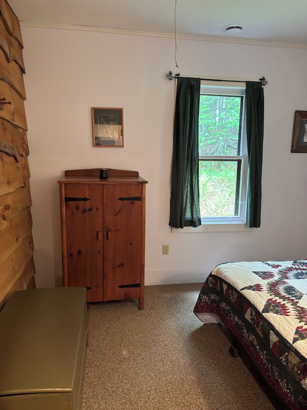 Une pièce avec une armoire en bois, un lit recouvert d’une courtepointe, une fenêtre avec des rideaux et un tableau encadré accroché au mur.