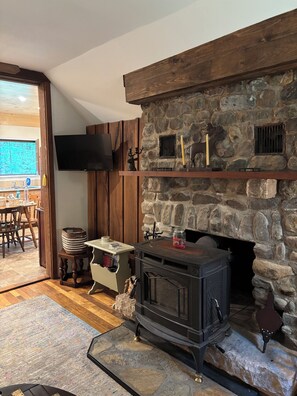 A cozy living room with a stone fireplace, wooden furniture, and a television mounted on the wall. - Quaint 2-bedroom cabin on Oxbow Lake perfect for relaxing Adirondack getaways (Lake Pleasant)