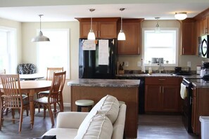 A kitchen with wooden cabinets, a black refrigerator, a dining area with a table and chairs, and pendant lighting. - Charming 3-bedroom cottage in lovely Vinalhaven perfect for relaxing (Vinalhaven)
