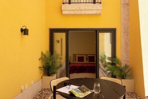 A yellow wall with a sliding glass door leading to a bedroom with a bed and red bedding. There is a table with chairs, a plant, and a glass of water. - Charming 2BR with Private Patio in São Bento (Lisbon)