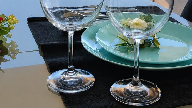 Una mesa con dos copas de vino y un plato con una flor, frente a una silla de mimbre y vistas al océano.