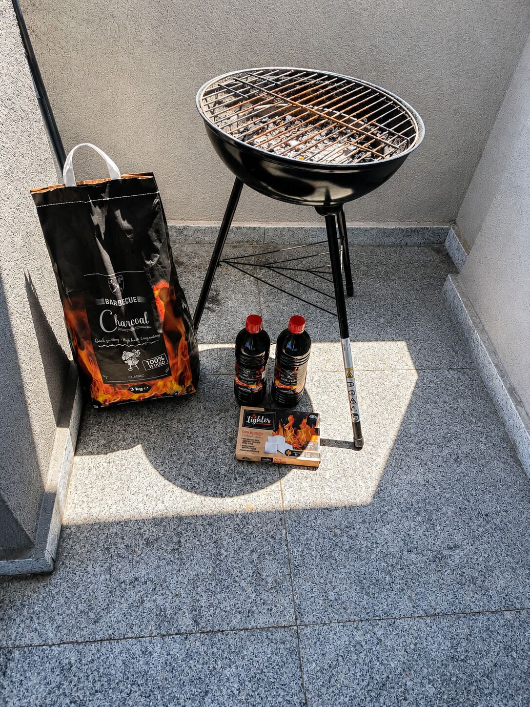 Ein Grill, Holzkohlebeutel und Feuerzeugflaschen auf einer gefliesten Terrasse.