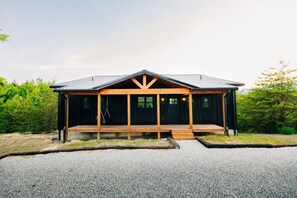 Uma cabana com varanda, entrada de cascalho e um fundo arborizado.