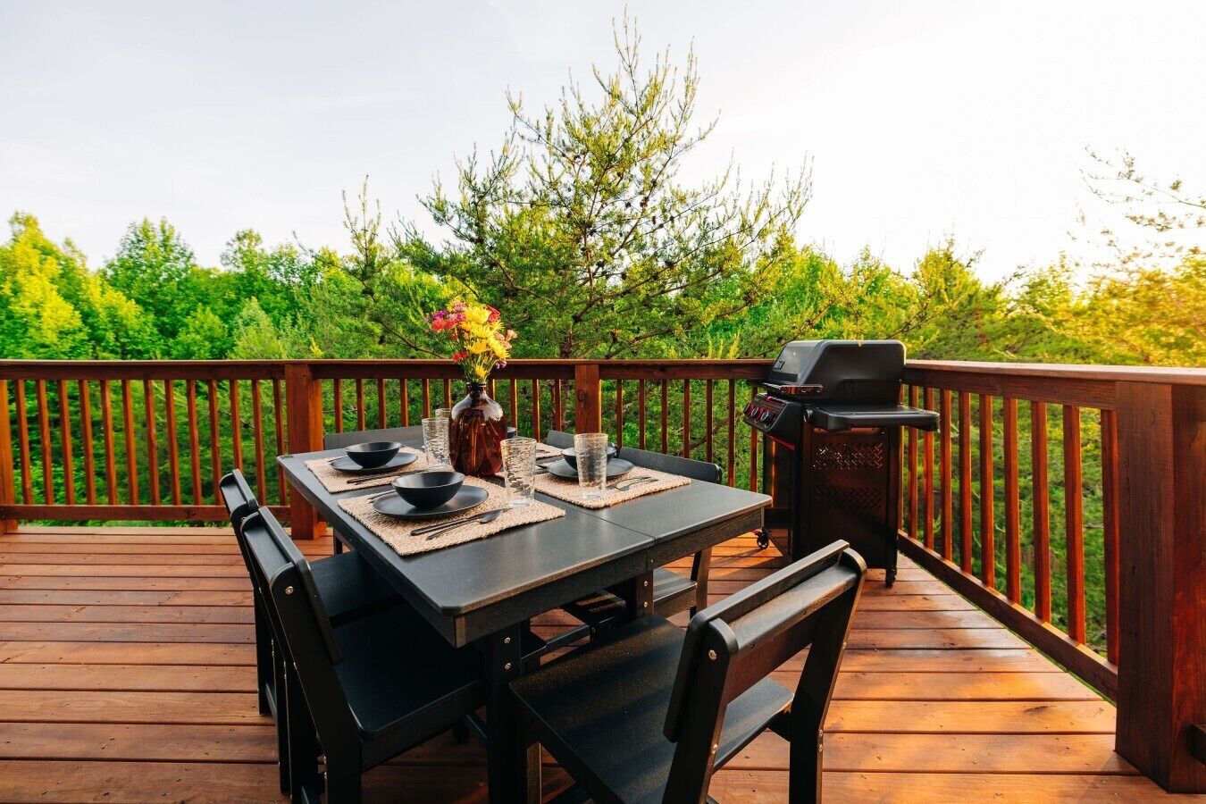 A wooden deck with a table set for dining, a grill, and a vase with flowers.
