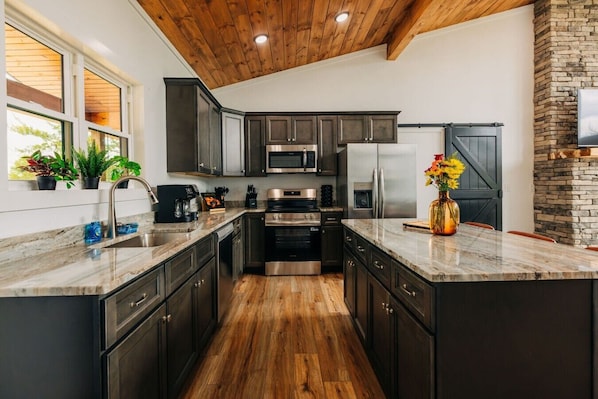 A modern kitchen with dark cabinets, stainless steel appliances, a wooden ceiling, and a stone fireplace. - Red River Gorge Cabin: Hot Tub, Fireplace + Deck (Zachariah)