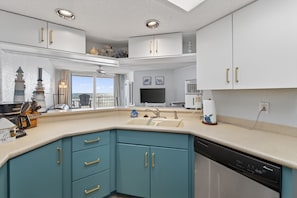 A modern kitchen with a dishwasher, a sink, and a window overlooking a balcony. - Sunrise Views & Sandy Toes – Beachfront Ponce Inlet Condo (Ponce Inlet)