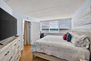 A bedroom with a bed, a TV, a dresser, a door, and a window with a view. - Sunrise Views & Sandy Toes – Beachfront Ponce Inlet Condo (Ponce Inlet)
