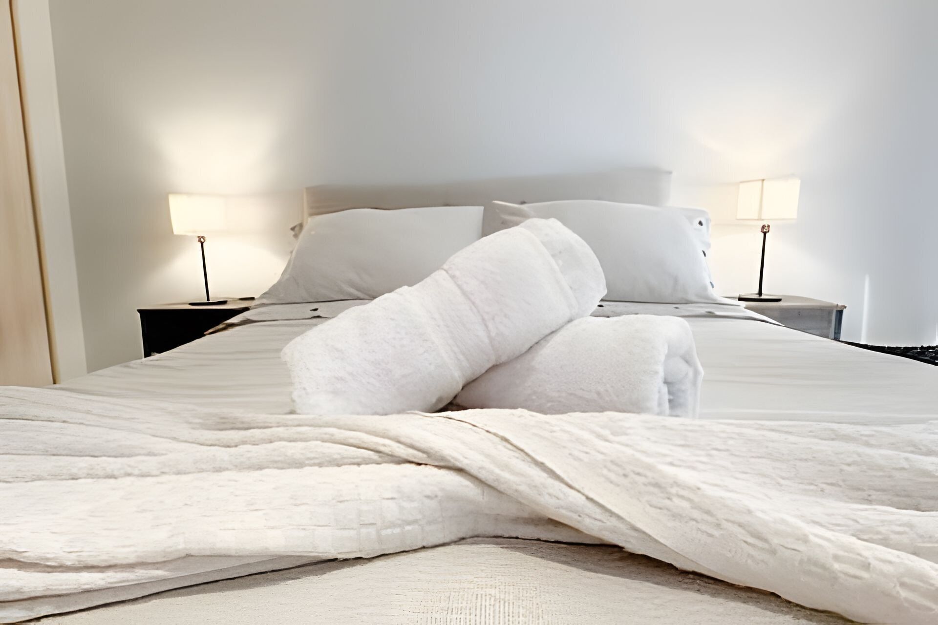 A neatly made bed with white linens and two pillows, a bedside table with a lamp, and a nightstand with a lamp.