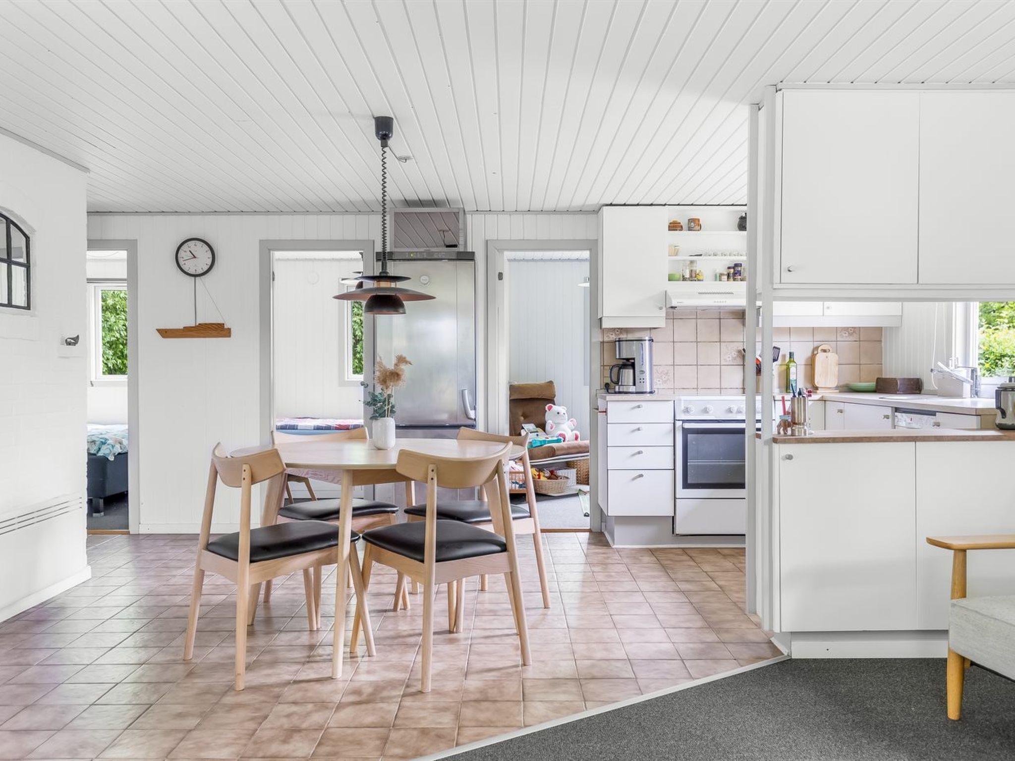 Une cuisine moderne avec des meubles blancs, un coin repas avec des chaises en bois et une horloge visible au mur.
