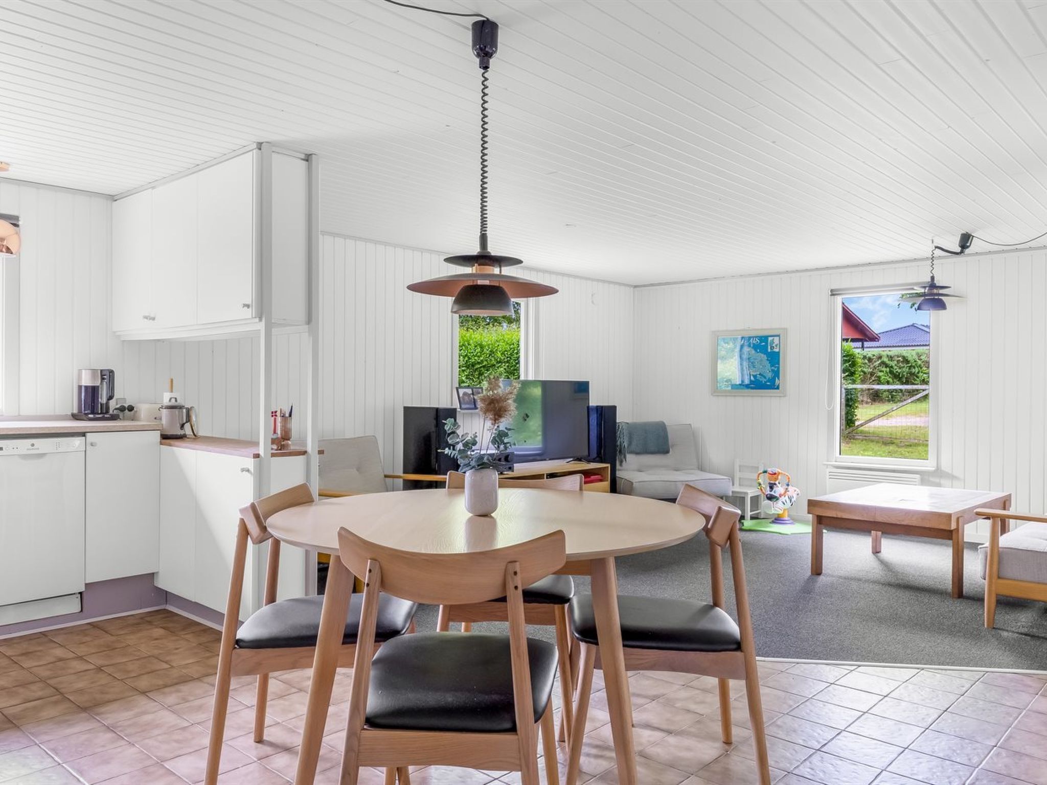 A modern kitchen with white cabinetry, a dining area with a round table and chairs, a television, and a view of greenery outside.