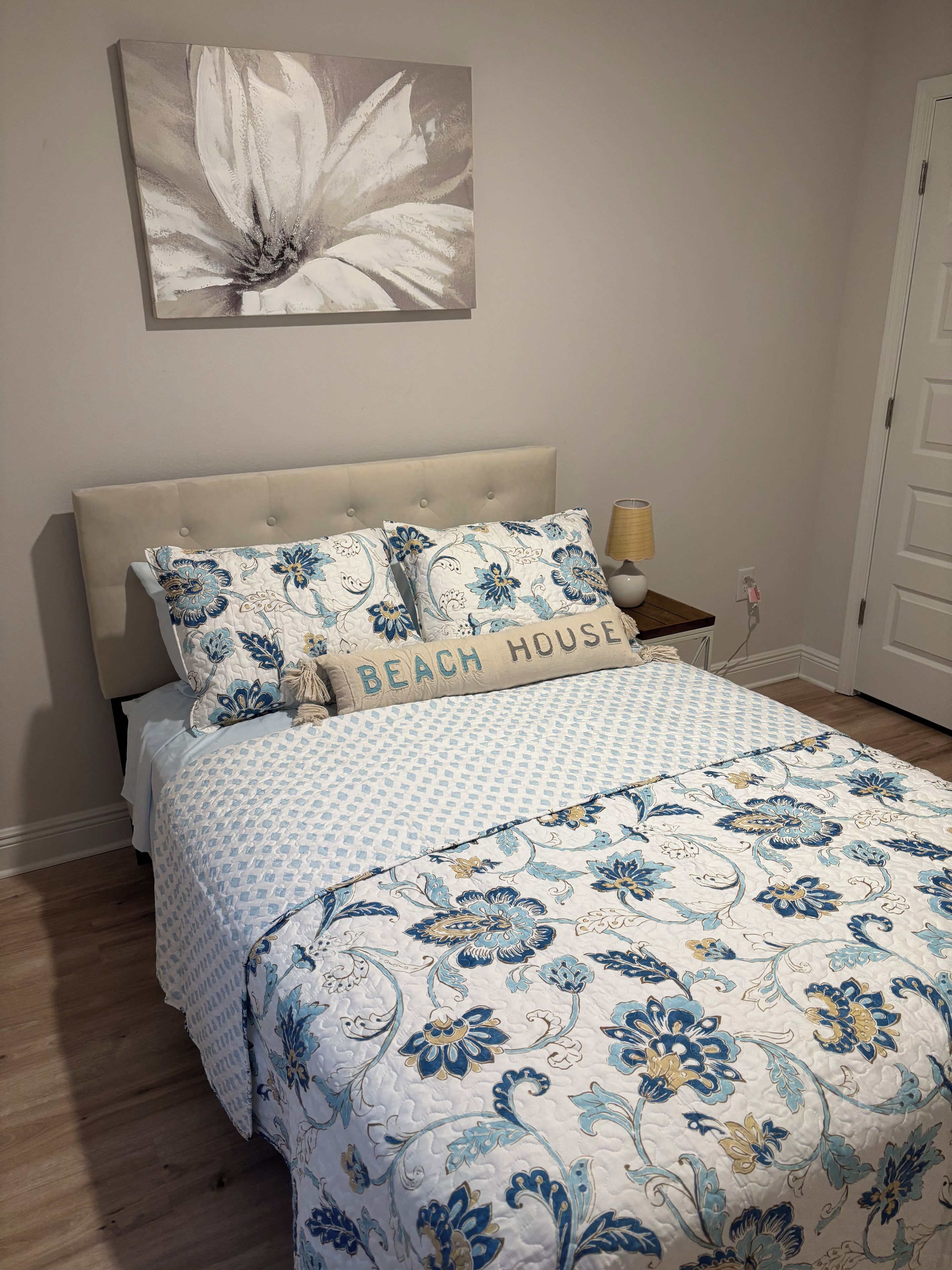 Um quarto com cama, mesa de cabeceira, abajur, obra de arte na parede e porta.