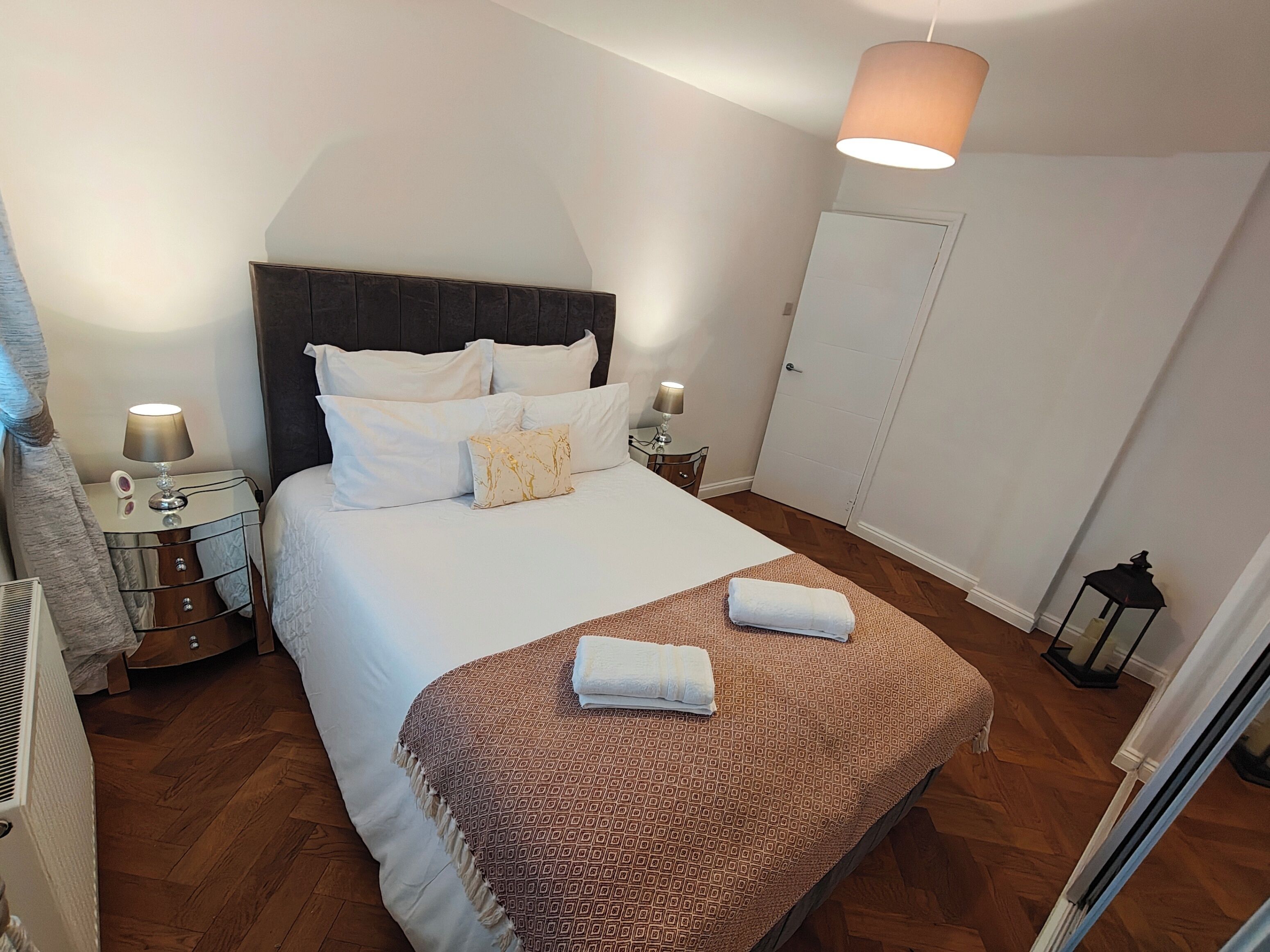 A neatly made bed with a patterned blanket, two bedside lamps, a mirror, and a wardrobe in a well-lit room with wooden flooring.