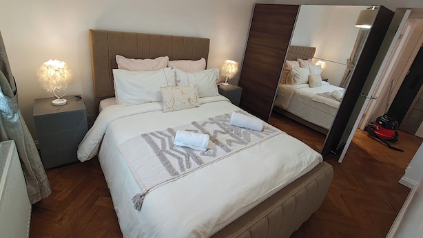 A bedroom with a large bed, two bedside tables with lamps, a mirror, and a vacuum cleaner. - Lovely Clydebank Apartment - 2 Bedrooms (Clydebank)
